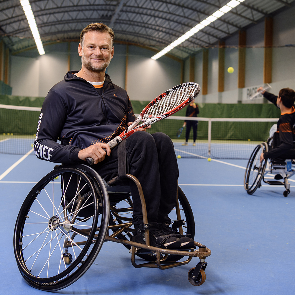 Porträttbild på Niclas Rodhborn, ledare inom rullstolstennis. Bilden är tagen i en tennishall med spelare i rullstol som spelar tennis bakom honom.