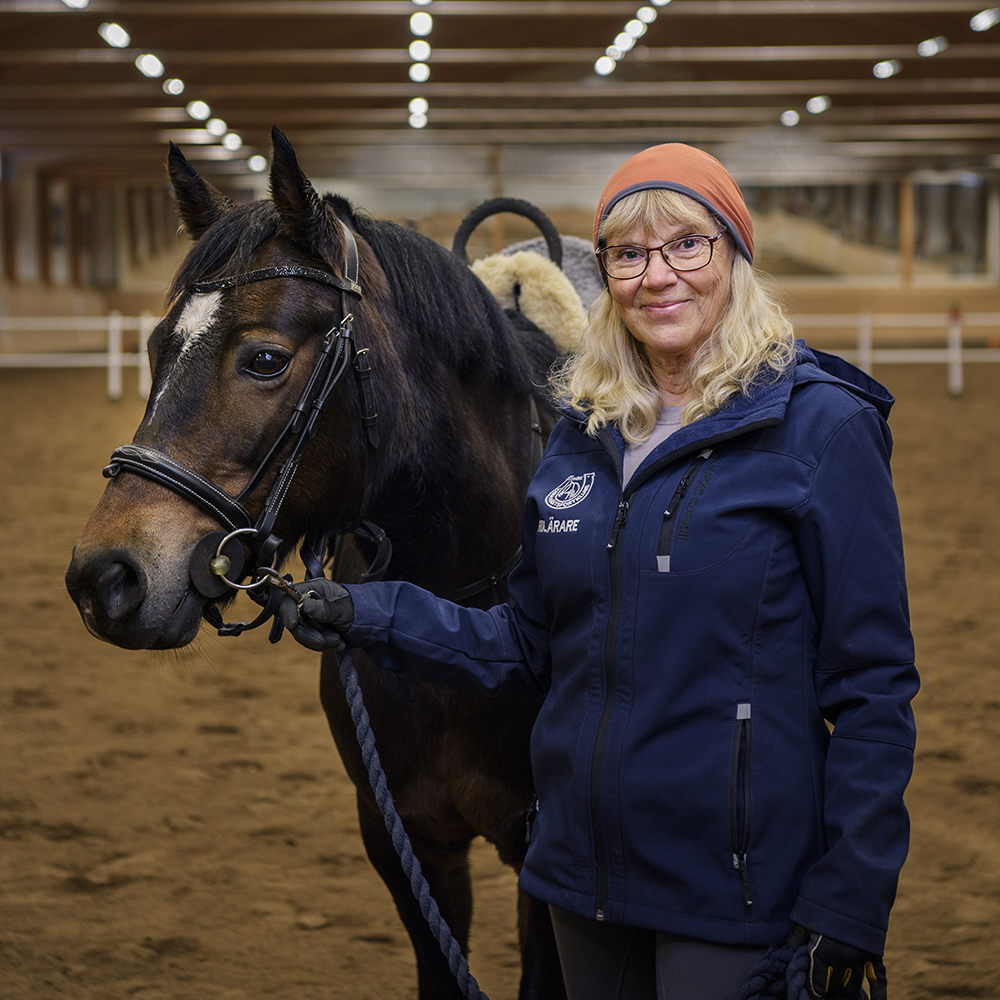 Porträttbild på Marita Matsson, ledare inom ridning. Marita har blå jacka, orange mössa och glasögon. Hon står i ett ridhus och bredvid en brun häst.