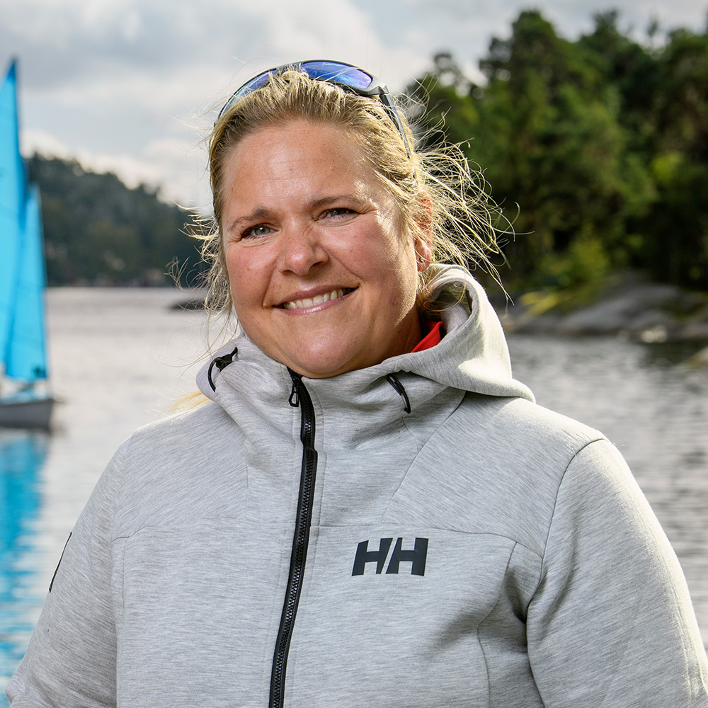 Porträttbild på Emma Hallén, ledare inom segling. Bilden är tagen utomhus vid vatten och i bakgrunden skymtar man en segelbåt.
