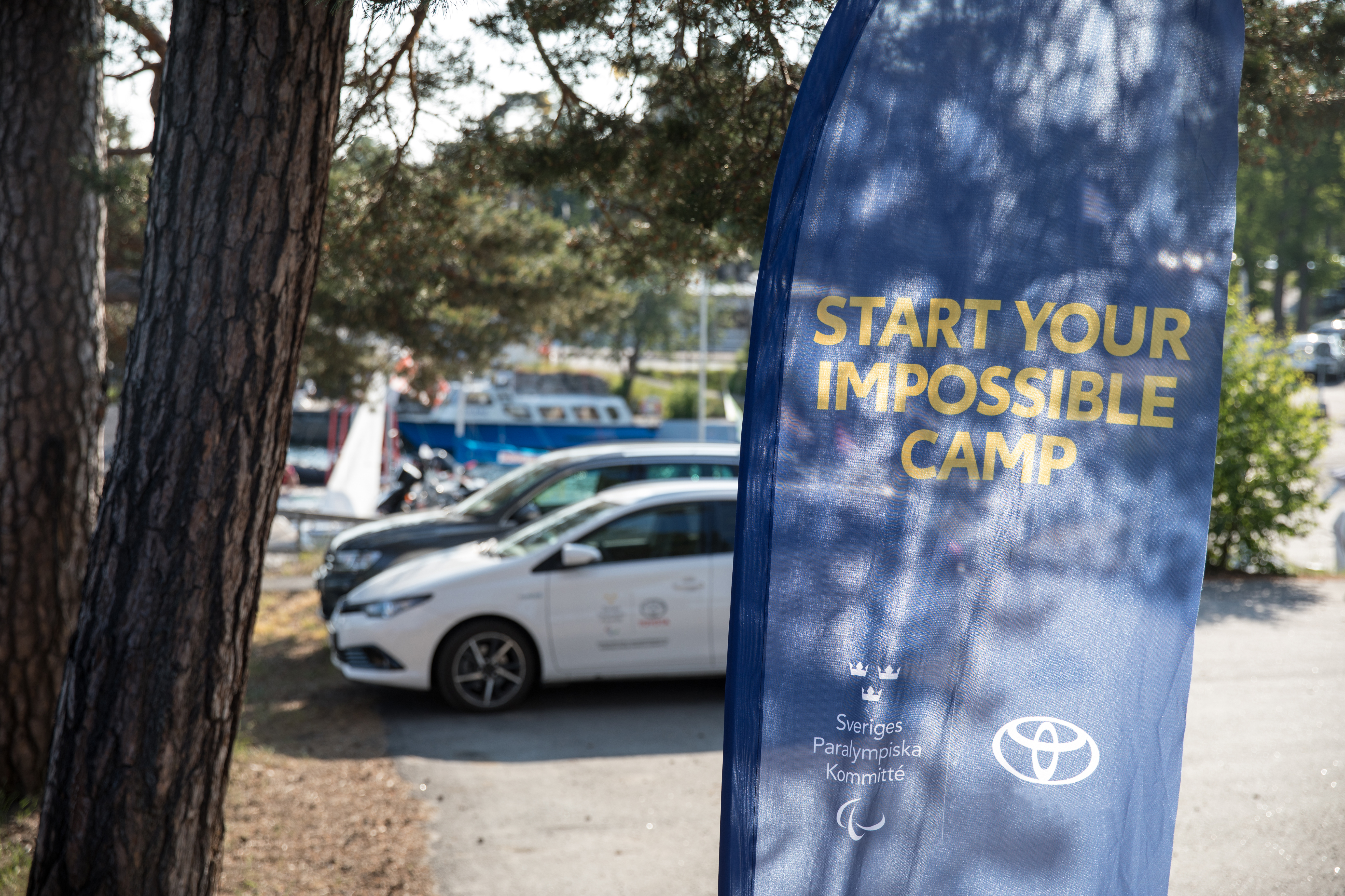 Utomhusmiljö. I förgrunden en flagga med texten Start Your Impossible Camp och logotypen för Sveriges Paralympiska Kommitté och Toyota. I bakgrunden en grusplan med en vit Toyota-bil parkerad.